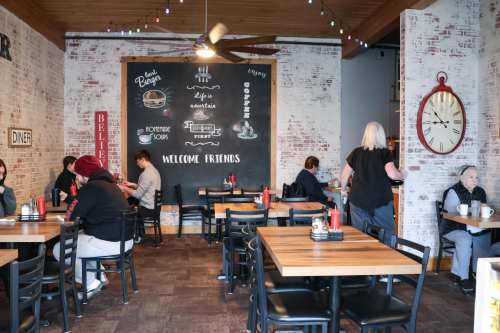 Cozy diner interior with wooden tables, chairs, and a chalkboard wall. Customers enjoy meals and coffee in a welcoming atmosphere.