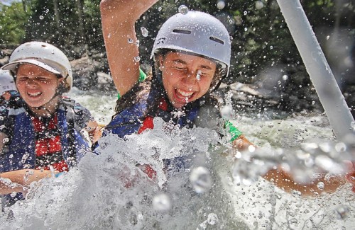 Two young girls joyfully rafting through splashing water, wearing helmets and life jackets, with smiles on their faces.