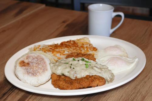 A plate of chicken fried steak with gravy, poached eggs, hash browns, and an English muffin, with a coffee cup in the background.