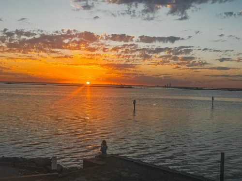 A serene sunset over a calm water body, with a person sitting on the shore, silhouetted against the vibrant sky.