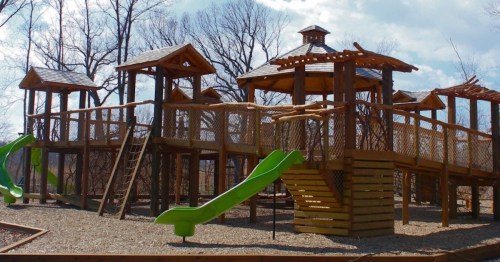 A wooden playground structure with slides, climbing areas, and a gazebo, surrounded by trees and a gravel surface.