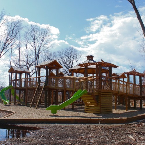A wooden playground with slides and climbing structures, surrounded by bare trees and a blue sky with clouds.