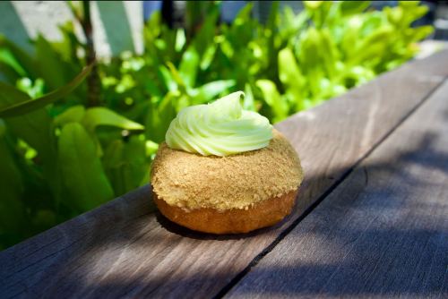 A cupcake with green frosting and a crumbly topping sits on a wooden surface, surrounded by lush green foliage.