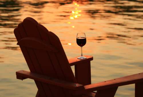 A glass of red wine rests on an Adirondack chair by a tranquil lake at sunset.