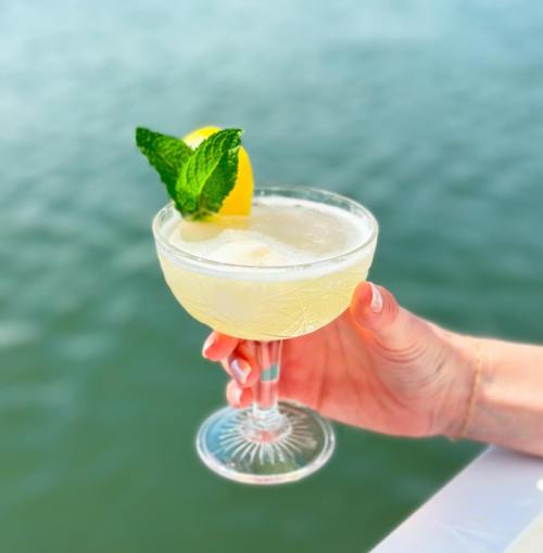 A hand holds a cocktail glass with a lemon slice and mint, set against a blurred water background.