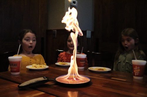 Three children watch in awe as a tall flame erupts from a dish on a table, surrounded by plates of food.