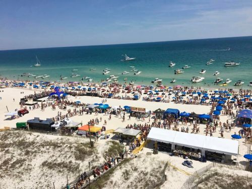 A crowded beach scene with people, colorful umbrellas, and boats on the water under a clear blue sky.