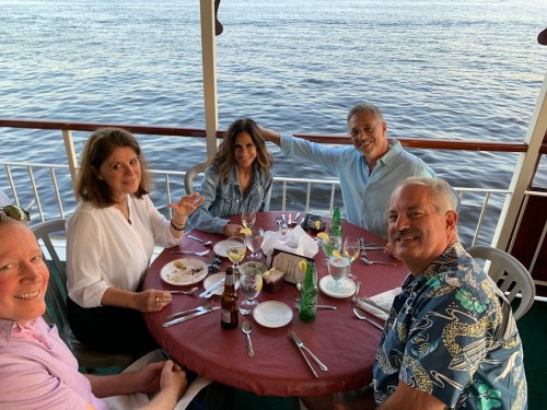 A group of five people enjoying a meal at a table by the water, smiling and engaging with each other.