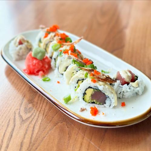 A plate of sushi rolls with avocado, tuna, and toppings, garnished with wasabi and pickled ginger on a wooden table.