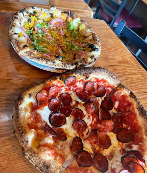 A close-up of two pizzas on a wooden table: one with pepperoni and cheese, the other topped with vegetables and herbs.