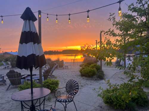 A serene beach scene at sunset, featuring a table, chairs, and string lights illuminating the tranquil atmosphere.