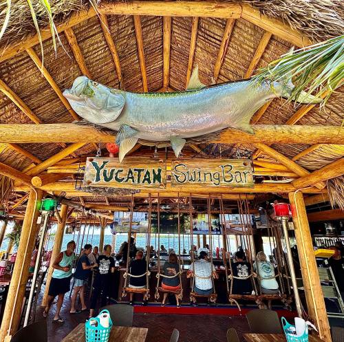 A tropical bar with a thatched roof, featuring a large fish decoration and patrons sitting on swings at the counter.