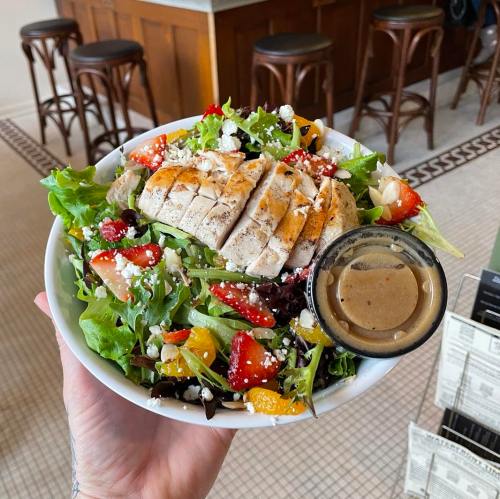 A hand holding a bowl of salad topped with grilled chicken, mixed greens, strawberries, and a side of dressing.