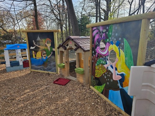 Colorful playground scene with two large murals and a playhouse, surrounded by wood chips and trees.