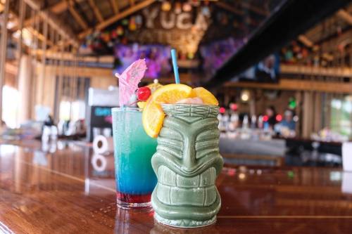 Two colorful cocktails on a wooden bar, one in a tiki mug with fruit garnishes, set in a vibrant tropical venue.