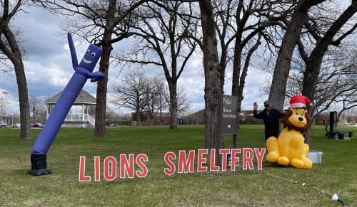 A blue inflatable tube man and a lion mascot stand beside a sign reading "LIONS SMELTFRY" in a park setting.