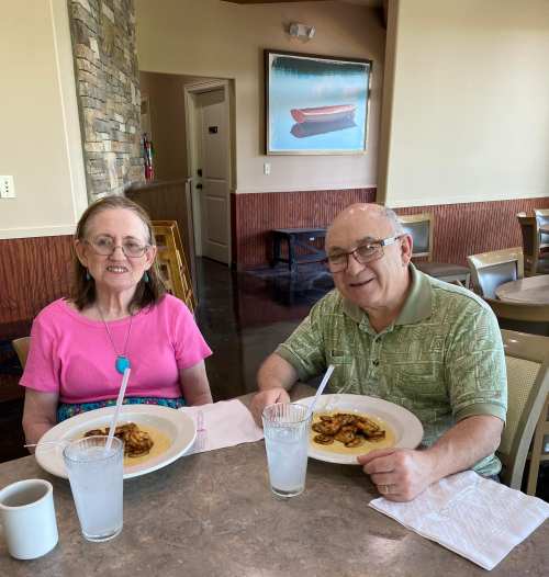 An elderly couple sits at a table in a restaurant, smiling with plates of food and glasses of water in front of them.