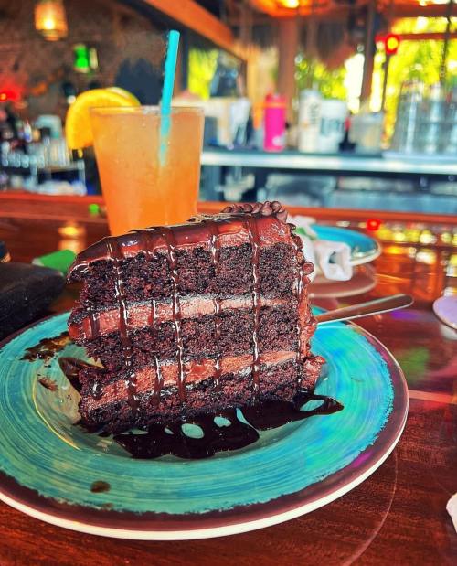 A slice of rich chocolate cake drizzled with sauce, next to a colorful drink on a wooden table.