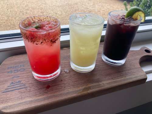 Three colorful cocktails on a wooden tray: a red drink with a rim, a light yellow drink, and a dark beverage with a lime slice.