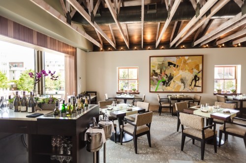 A modern restaurant interior with wooden beams, elegant tables, and a large abstract painting on the wall.