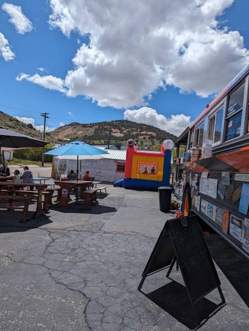 A food truck and picnic area with a bouncy castle under a blue sky with fluffy clouds and mountains in the background.