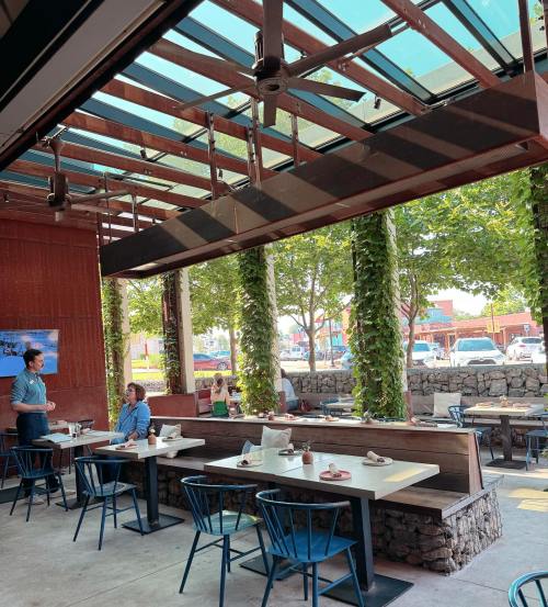 A modern restaurant interior with large windows, greenery, and diners at tables, creating a bright and inviting atmosphere.