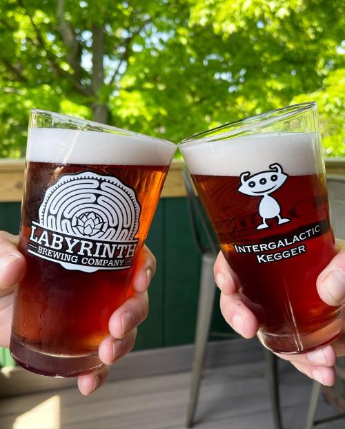 Two glasses of amber beer clink together, featuring logos for Labyrinth Brewing Company and Intergalactic Kegger.