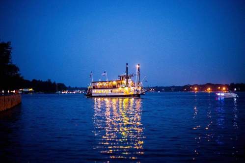 A lit steamboat glides across a calm lake at dusk, reflecting lights on the water's surface.