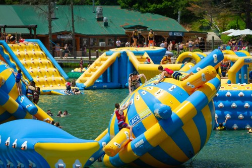 Colorful inflatable water park with people climbing and playing on structures in a lake, surrounded by trees and a building.