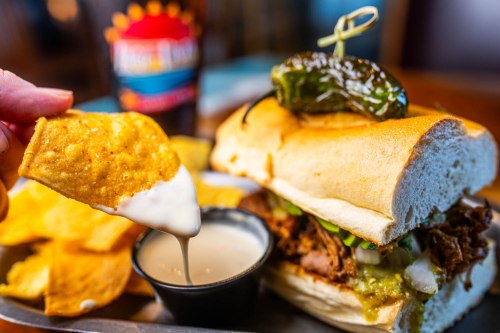 A hand dips a chip into creamy sauce beside a sandwich filled with pulled meat, topped with a pepper and served with chips.
