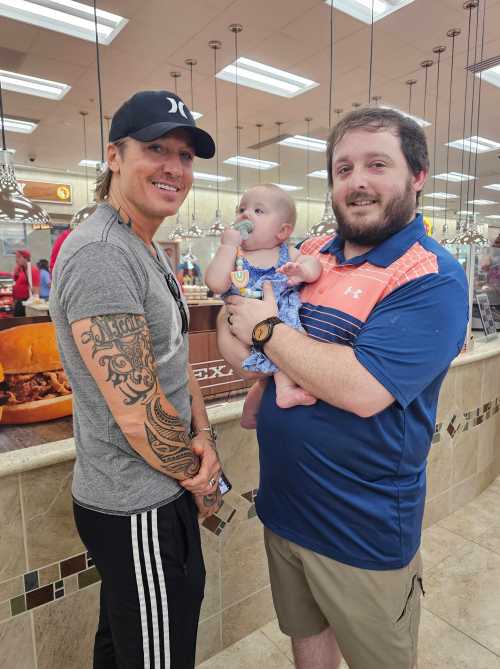 Two men stand in a restaurant, one holding a baby. They are smiling and chatting in a casual setting.