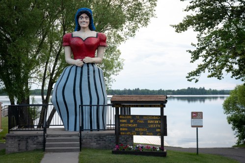A large statue of a woman in a red top and striped skirt stands by a lake, with trees and a sign nearby.