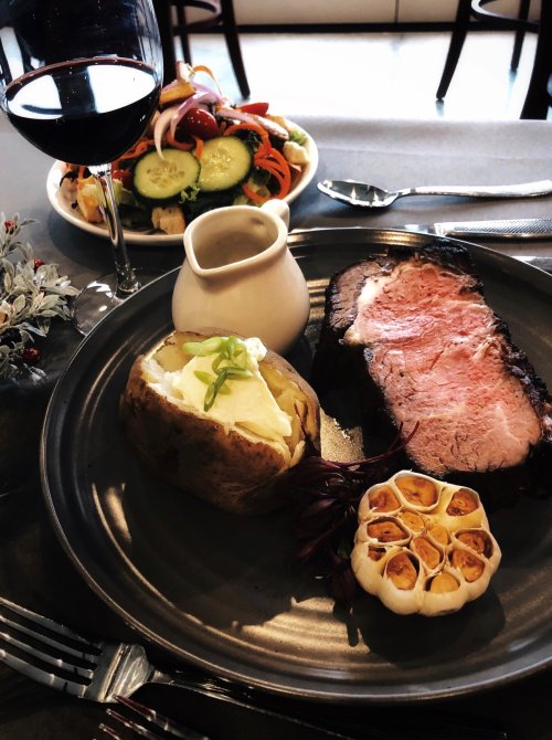 A plate with sliced roast beef, a baked potato with sour cream, roasted garlic, and a side salad, accompanied by red wine.