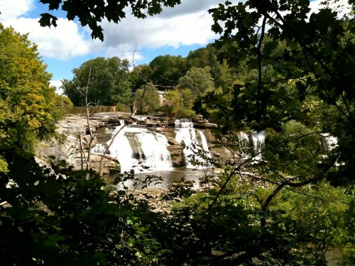 A serene waterfall cascades over rocks, surrounded by lush greenery and trees under a partly cloudy sky.