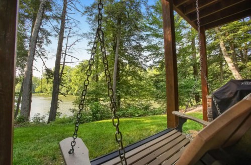 A wooden swing chair on a porch overlooking a serene river and lush green trees.