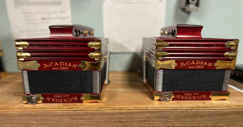Two red and gold Acadian accordions sit side by side on a wooden surface, showcasing their intricate design.