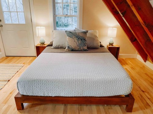 Cozy bedroom with a wooden bed, decorative pillows, two lamps, and a window overlooking a wooded area.