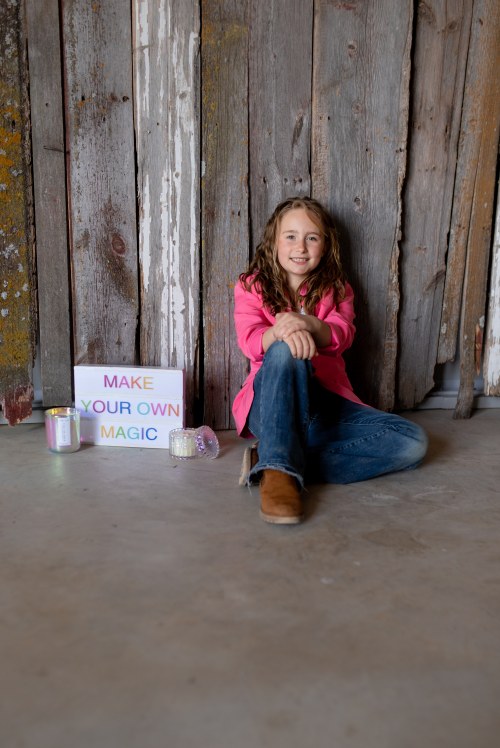A girl in a pink jacket sits on the floor next to a sign that says "MAKE YOUR OWN MAGIC," with rustic wooden walls behind her.