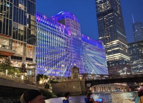 A vibrant cityscape at night featuring a building illuminated with colorful lights along a river.