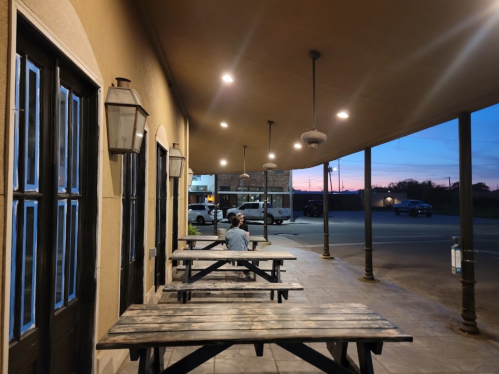 A person sits alone at a picnic table outside a building, with a sunset visible in the background.