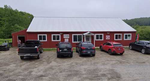 A red barn-style store with a white roof, surrounded by parked cars on a gravel lot, set in a green landscape.