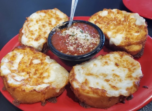 Four cheesy bread slices arranged around a bowl of marinara sauce, garnished with grated cheese, on a red plate.