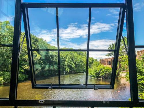 A large window opens to a view of a river surrounded by lush greenery and a blue sky with scattered clouds.