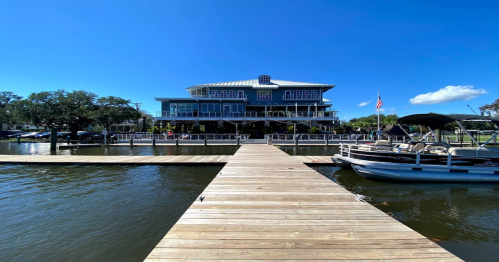 A wooden dock leads to a large blue building by the water, with boats moored nearby and a clear blue sky above.