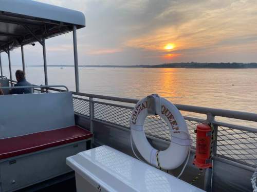 A sunset over calm waters viewed from a boat, with a lifebuoy and seating visible on the deck.