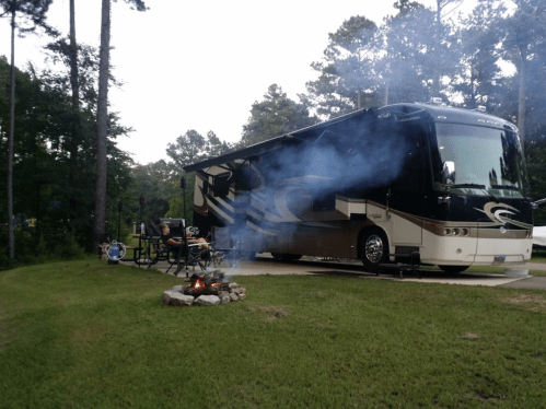An RV parked on a grassy area with a campfire nearby, surrounded by trees and smoke rising into the air.