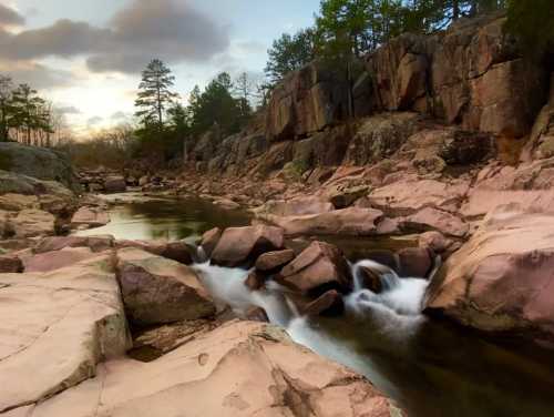 A serene river flows over smooth rocks, surrounded by towering cliffs and trees under a cloudy sky at sunset.