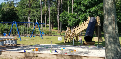 A playground featuring swings, a slide, and a sand area with colorful toys, surrounded by trees.