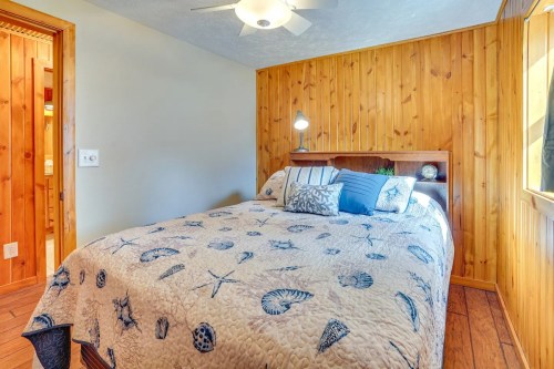 Cozy bedroom with wooden walls, a patterned bedspread featuring seashells, and a ceiling fan.