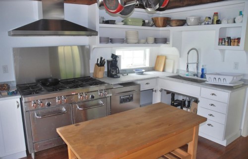 A modern kitchen featuring stainless steel appliances, a wooden table, and open shelving with dishes and utensils.